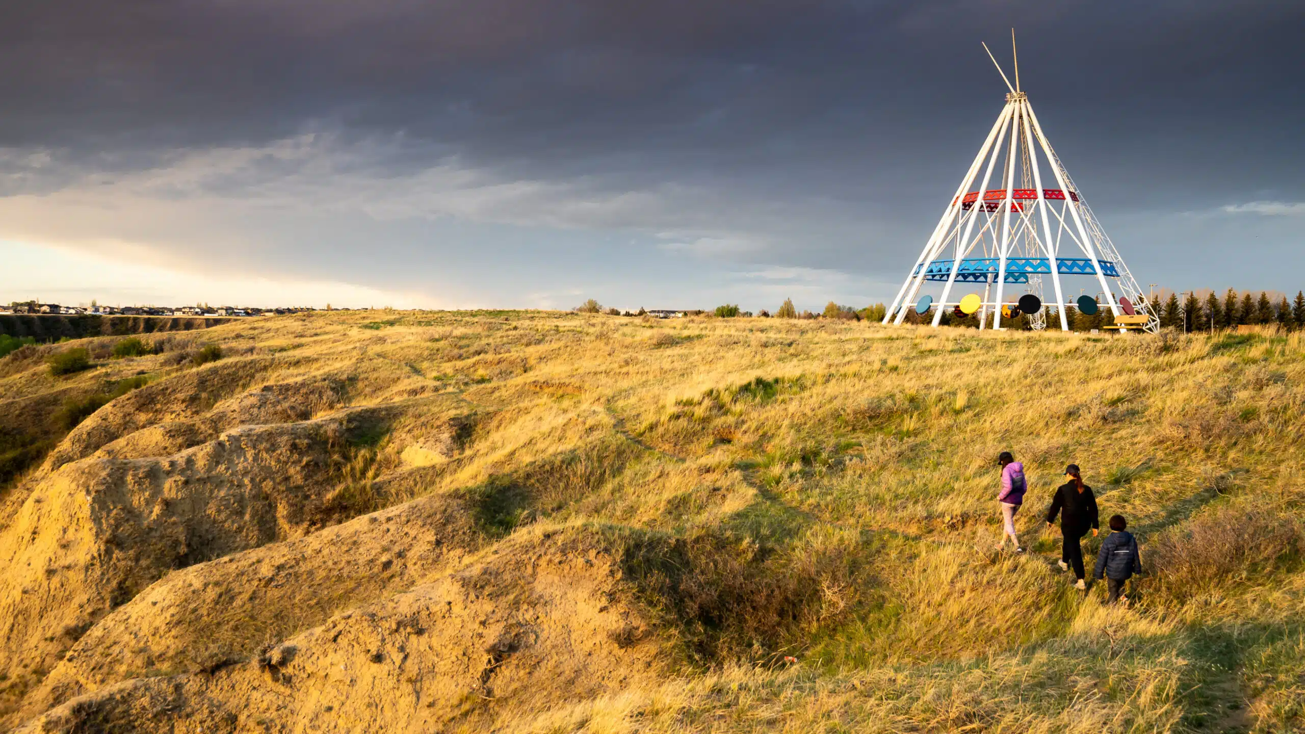 Three people walk up a grassy hill toward a large white metal tipi structure.